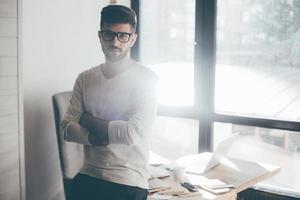 empresário confiante. jovem confiante em óculos, mantendo os braços cruzados e olhando para a câmera enquanto se inclina na mesa no escritório foto