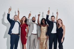 grupo de pessoas bonitas em trajes formais brindando com champanhe e sorrindo em pé contra um fundo cinza foto