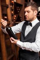 este vinho é perfeito. sommelier masculino confiante examinando garrafa de vinho em pé perto da prateleira de vinho foto