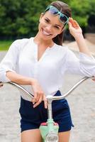 beleza na moto. bela jovem sorridente ajustando seus óculos de sol enquanto se inclina em sua bicicleta vintage no parque foto