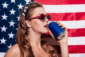 tomando um gole. mulher jovem e bonita usando óculos escuros e bebendo de lata em pé contra a bandeira nacional americana foto