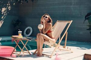 mulher jovem e atraente de biquíni relaxando e desfrutando de um coquetel refrescante enquanto toma sol à beira da piscina ao ar livre foto