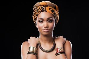 Rainha africana. linda mulher africana usando um lenço na cabeça e tocando seus ombros em pé contra um fundo preto foto