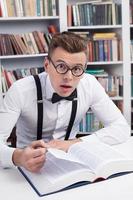 nerd chocado. jovem surpreso de camisa e gravata borboleta lendo um livro na biblioteca e olhando para a câmera foto