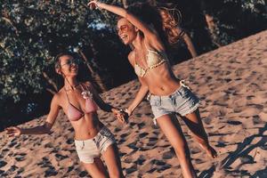 hora de diversão. duas mulheres jovens atraentes em shorts e trajes de banho sorrindo enquanto correm na praia foto