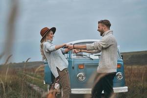 sentindo brincalhão. lindo casal jovem de mãos dadas e sorrindo em pé perto da mini van azul estilo retrô foto