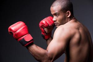 treinando suas habilidades de boxe. Vista lateral do jovem africano sem camisa em luvas de boxe exercitando em pé contra um fundo cinza foto