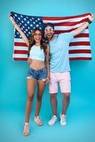 comprimento total do belo casal jovem carregando a bandeira americana e sorrindo em pé contra um fundo azul foto