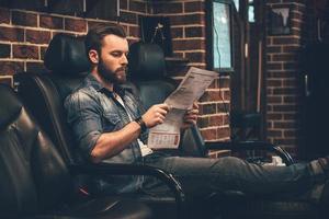 aguardando agendamento. bonito jovem barbudo lendo jornal enquanto está sentado em uma cadeira confortável na barbearia foto