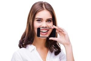 sorriso sincero. mulheres bonitas segurando o celular contra a boca e sorrindo em pé contra um fundo cinza foto