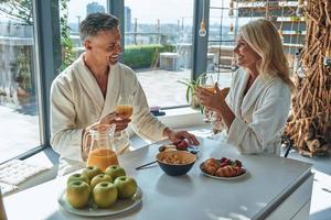 lindo casal maduro em roupões de banho tomando café da manhã juntos enquanto passa o tempo na cozinha doméstica foto