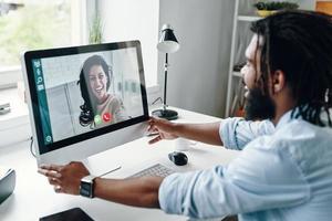 feliz jovem africano na camisa falando com uma mulher sorridente usando computador enquanto está sentado dentro de casa foto