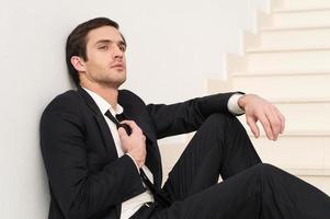 cansado do trabalho. Vista lateral do jovem pensativo em trajes formais, ajustando sua gravata enquanto está sentado na escada foto