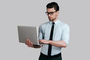usando laptop. jovem bonito de camisa branca e gravata trabalhando no laptop em pé contra um fundo cinza foto