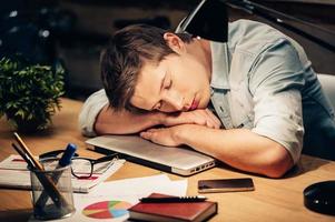 muito trabalho. jovem dormindo em seu local de trabalho enquanto inclina a cabeça no laptop foto