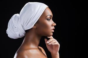 retrato da beleza. Vista lateral da bela mulher africana usando um lenço na cabeça e segurando a mão no queixo em pé contra um fundo preto foto