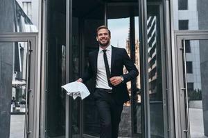 bonito jovem empresário de terno completo sorrindo e segurando o documento enquanto caminhava ao ar livre foto