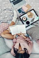 vista superior da bela jovem de pijama lendo livro e desfrutando de café da manhã enquanto descansa na cama em casa foto