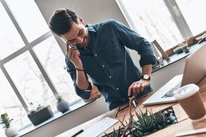 conversa importante. jovem bonito falando no telefone inteligente e sorrindo enquanto trabalhava no escritório foto