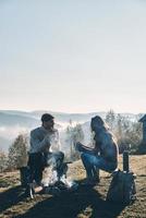 manhã tranquila. lindo casal jovem tomando café da manhã enquanto está sentado junto à fogueira nas montanhas foto