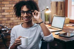 pausa para o café. alegre jovem africano segurando a xícara de café e ajustando seus óculos com sorriso enquanto está sentado ao lado de seu local de trabalho foto