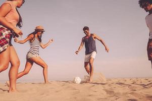 diversão de praia. grupo de jovens alegres brincando com bola de futebol na praia com mar ao fundo foto