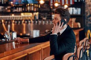 jovem bonito de terno completo falando no telefone inteligente enquanto está sentado no balcão do bar no restaurante foto