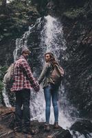 ficar longe de tudo... vista traseira de corpo inteiro do jovem casal de mãos dadas em pé perto da cachoeira foto