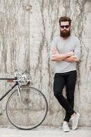 encantador bonito. comprimento total de jovem barbudo mantendo os braços cruzados e olhando para a câmera em pé perto de sua bicicleta contra a parede de concreto foto