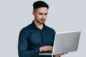 concentrado no trabalho. jovem bonito na camisa trabalhando no laptop em pé contra um fundo cinza foto