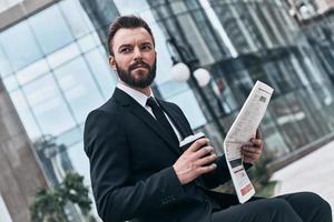 confiante e inteligente. jovem bonito de terno completo lendo um jornal enquanto está sentado ao ar livre foto