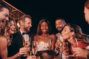 mulher jovem e bonita no vestido de cokctail soprando velas no bolo de aniversário enquanto passa o tempo na festa com os amigos foto