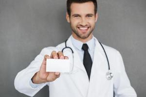 médico de família de confiança. sorridente jovem médico de uniforme branco esticando seu cartão de visita em pé contra um fundo cinza foto