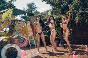 comprimento total de mulheres jovens atraentes em trajes de banho se divertindo com coquetéis refrescantes e sorrindo em pé à beira da piscina ao ar livre foto