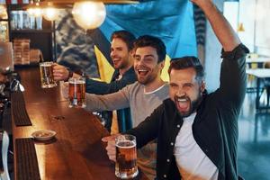 torcendo por jovens cobertos de bandeira ucraniana bebendo cerveja e assistindo jogo esportivo enquanto está sentado no pub foto