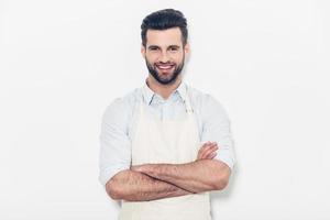 homem de avental. confiante jovem bonito no avental, mantendo os braços cruzados e sorrindo em pé contra o fundo branco foto