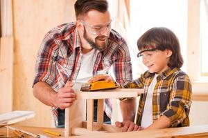 equipe de carpintaria. alegre jovem carpinteiro ajudando seu filho tosand cadeira de madeira na oficina foto