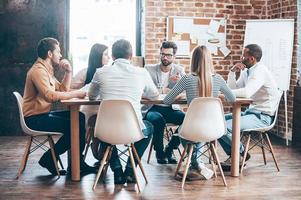 reunião matinal. grupo de seis jovens discutindo algo enquanto estão sentados à mesa no escritório juntos foto