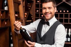 sommelier confiante. sommelier masculino confiante segurando a garrafa de vinho e sorrindo em pé perto da prateleira de vinho foto