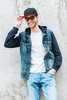 cara elegante. jovem bonito ajustando headwear e sorrindo em pé contra a parede de concreto foto