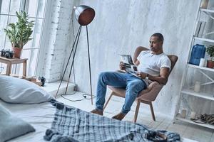 o que há de novo no mundo jovem africano bonito lendo jornal enquanto está sentado na poltrona em casa foto