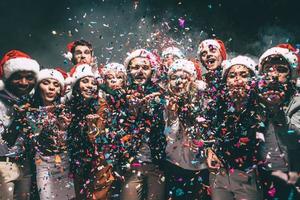 diversão colorida. grupo de jovens bonitos em chapéus de Papai Noel soprando confetes coloridos e parecendo feliz foto