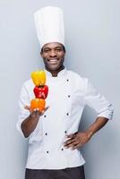 culinária colorida. confiante jovem chef africano em uniforme branco segurando pimentões multicoloridos e olhando para a câmera com sorriso em pé contra um fundo cinza foto