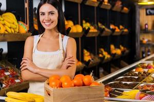 bem-vindo ao paraíso das frutas bela jovem de avental mantendo os braços cruzados e sorrindo em pé na mercearia com variedade de frutas ao fundo foto