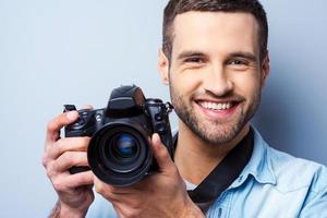 sorria para uma câmera jovem bonito segurando a câmera digital e sorrindo em pé contra um fundo cinza foto