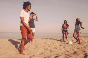 se divertindo na praia. grupo de jovens alegres brincando com bola de futebol na praia com mar ao fundo foto