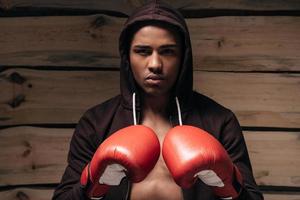 pronto para lutar. confiante jovem africano em camisa com capuz e luvas de boxe, olhando para a câmera em pé contra um fundo de madeira foto