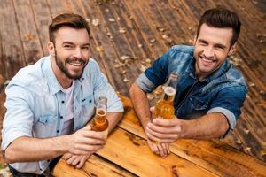 aplaude a vista superior de dois jovens alegres esticando garrafas com cerveja e olhando para a câmera em pé ao ar livre