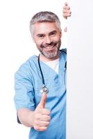 cirurgião de sucesso. cirurgião maduro confiante olhando para fora do cartaz branco e mostrando o polegar para cima em pé isolado no branco foto