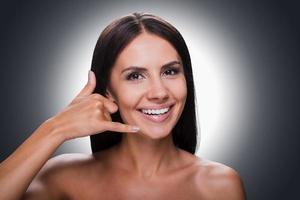 esperando seu retrato de chamada de sorridente jovem sem camisa olhando para a câmera e gesticulando o celular perto do rosto em pé contra um fundo cinza foto